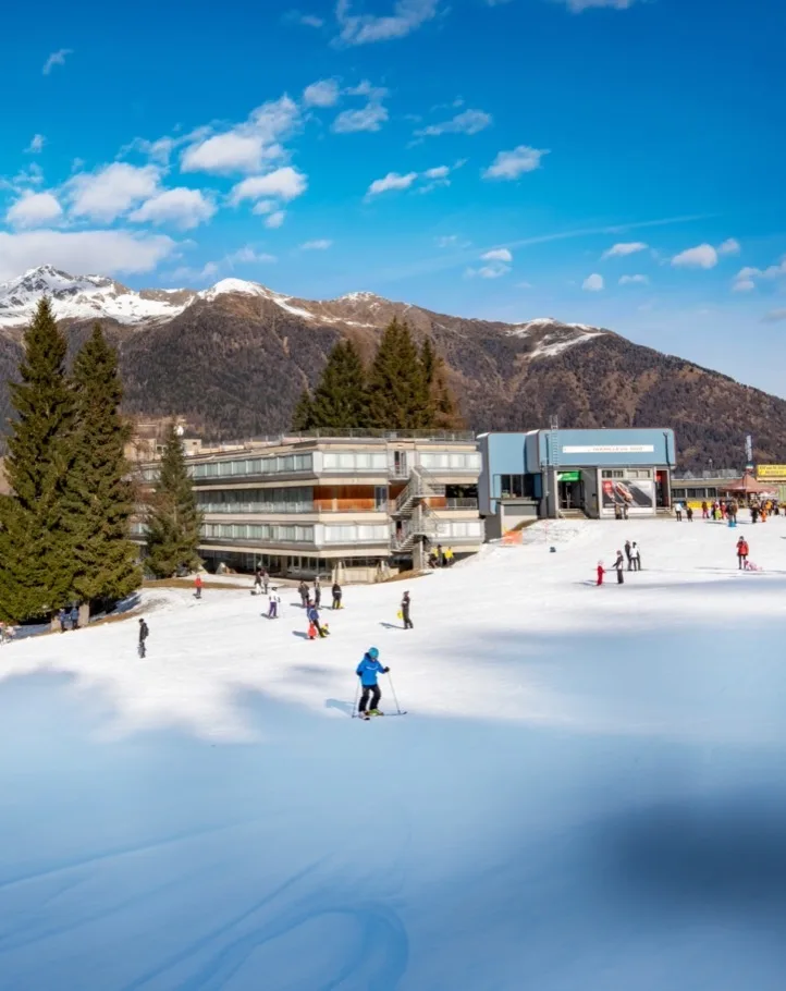 Hotel ai piedi delle piste con sciatori e montagne sullo sfondo in giornata di sole