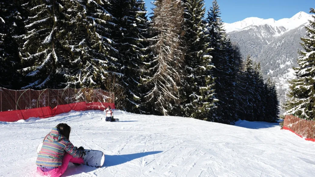 Bambina con snowboard seduta sulla pista da sci circondata da abeti innevati