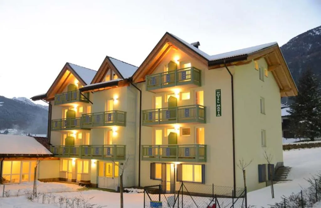 Facciata dell'Hotel Maria con balconi verdi e luci accese al tramonto sulla neve